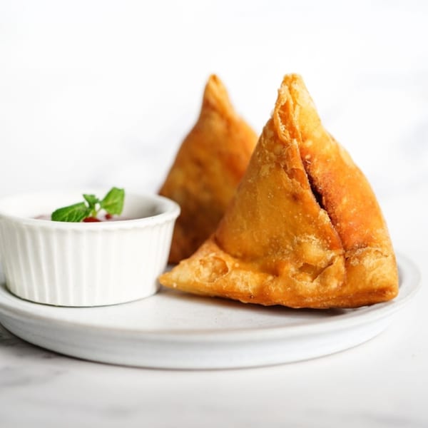 Two golden-brown samosas and a small white bowl of chutney with a mint garnish on a white plate.