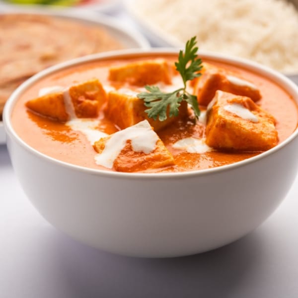A white bowl of paneer butter masala, garnished with cream and cilantro, with rice and naan in the background.