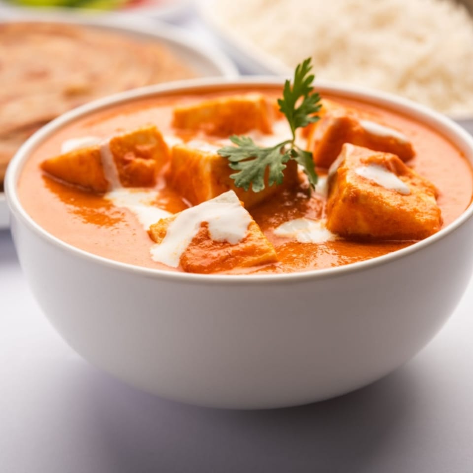 A white bowl of paneer butter masala, garnished with cream and cilantro, with rice and naan in the background.